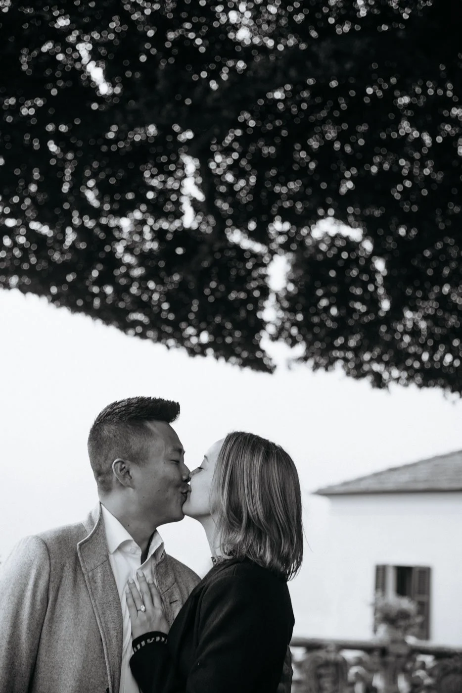 A couple sharing a kiss outdoors under a large, bokeh-blurred tree in black and white.