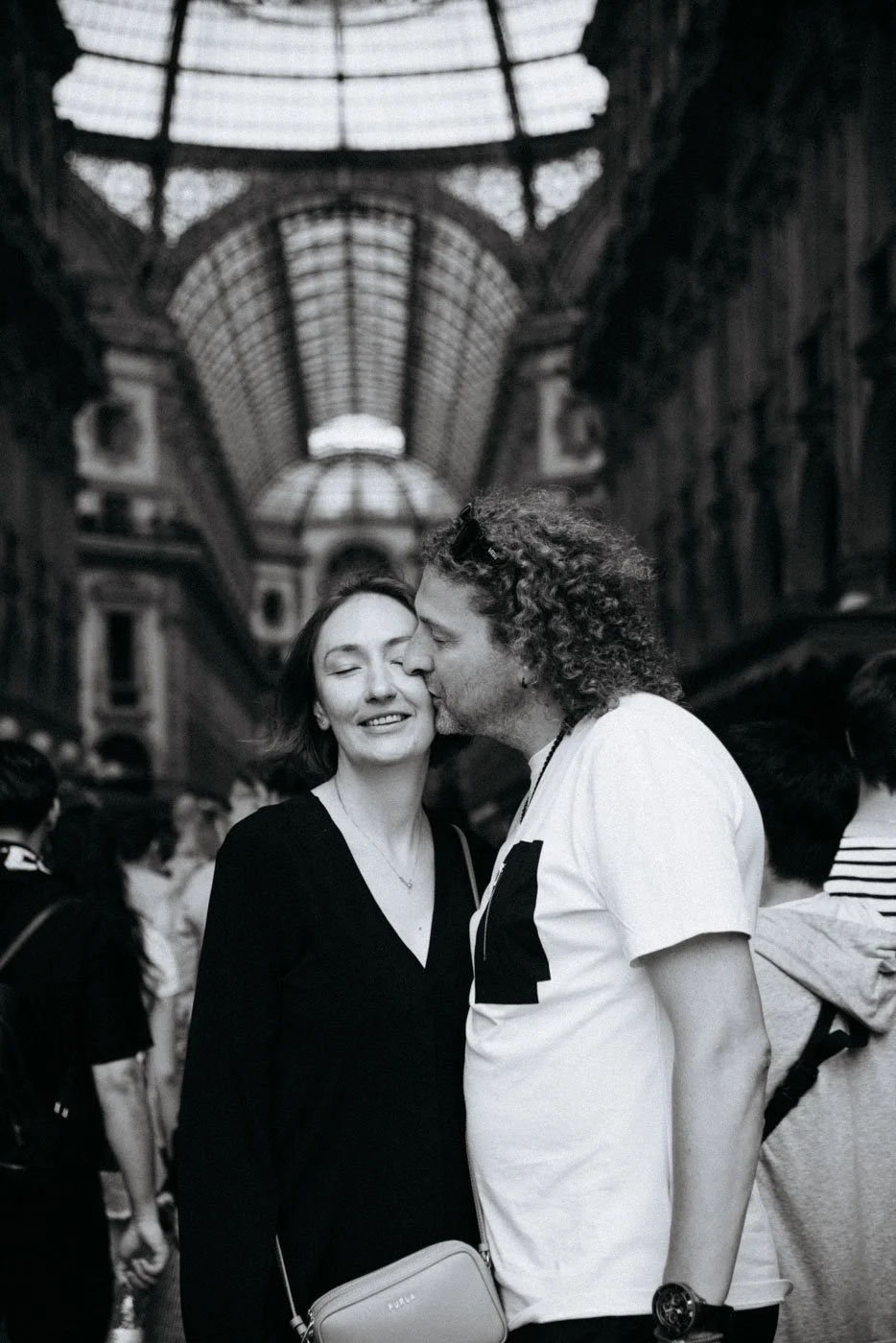 A man and woman in a crowded indoor setting, with the man kissing the woman's cheek; the woman smiling with eyes closed, wearing a black top and carrying a light-colored purse. The scene is in a large, glass-domed shopping arcade or mall.