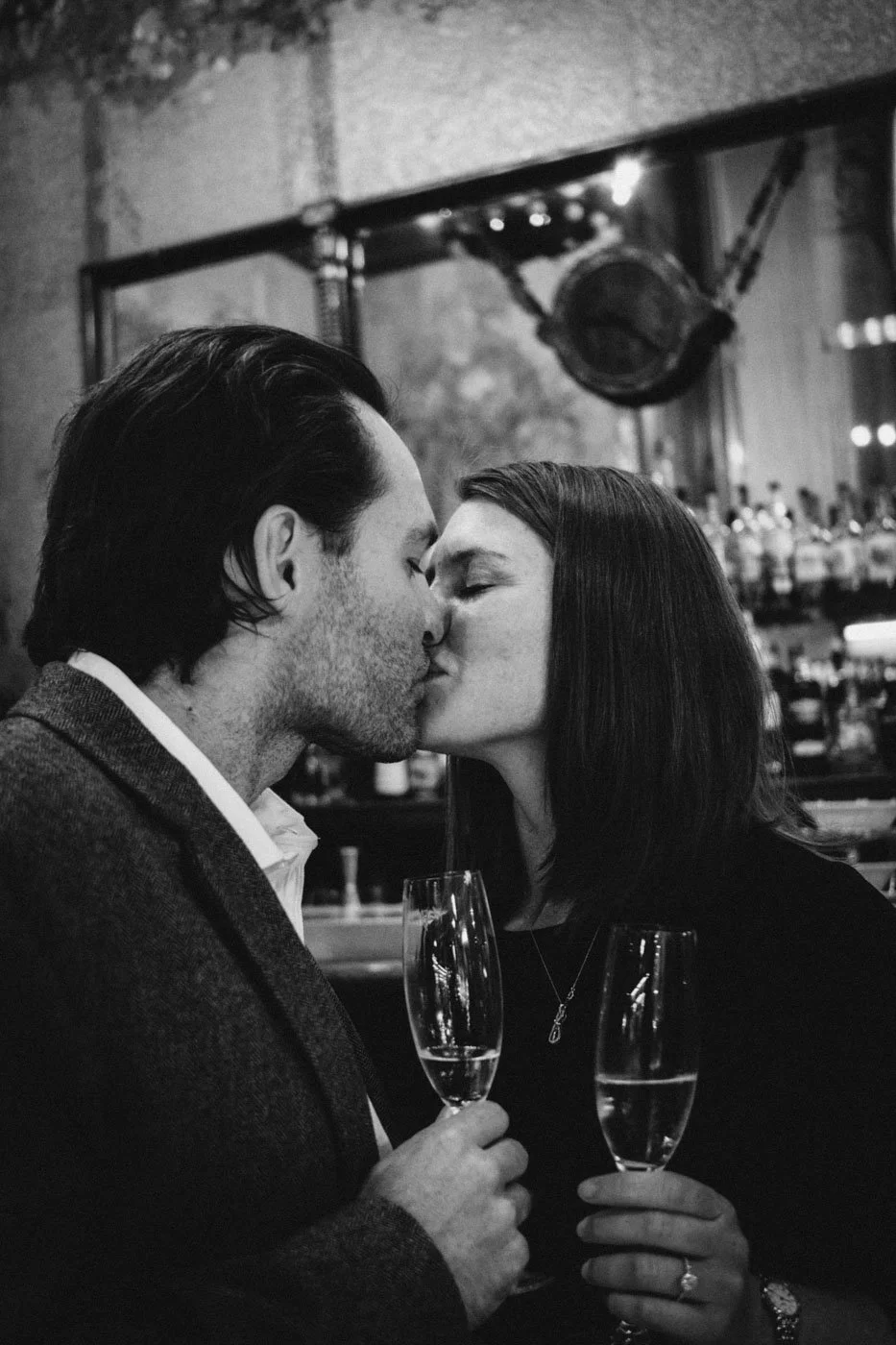 A black and white photo of a man and woman kissing each other at a bar, holding champagne glasses.