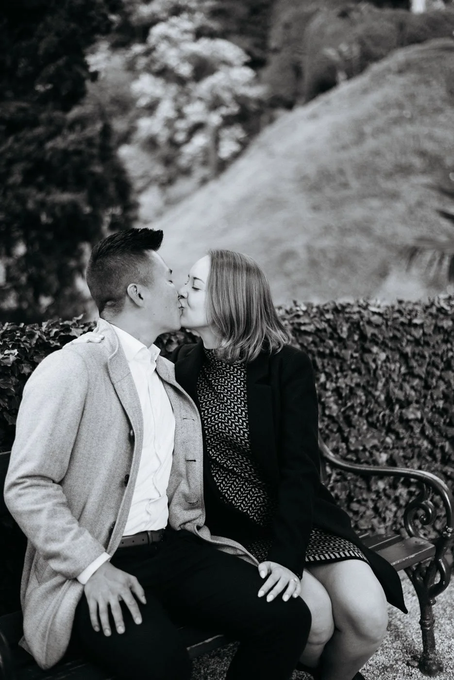 A black and white photo of a couple sitting on a bench, sharing a kiss outdoors, with trees and foliage in the background.