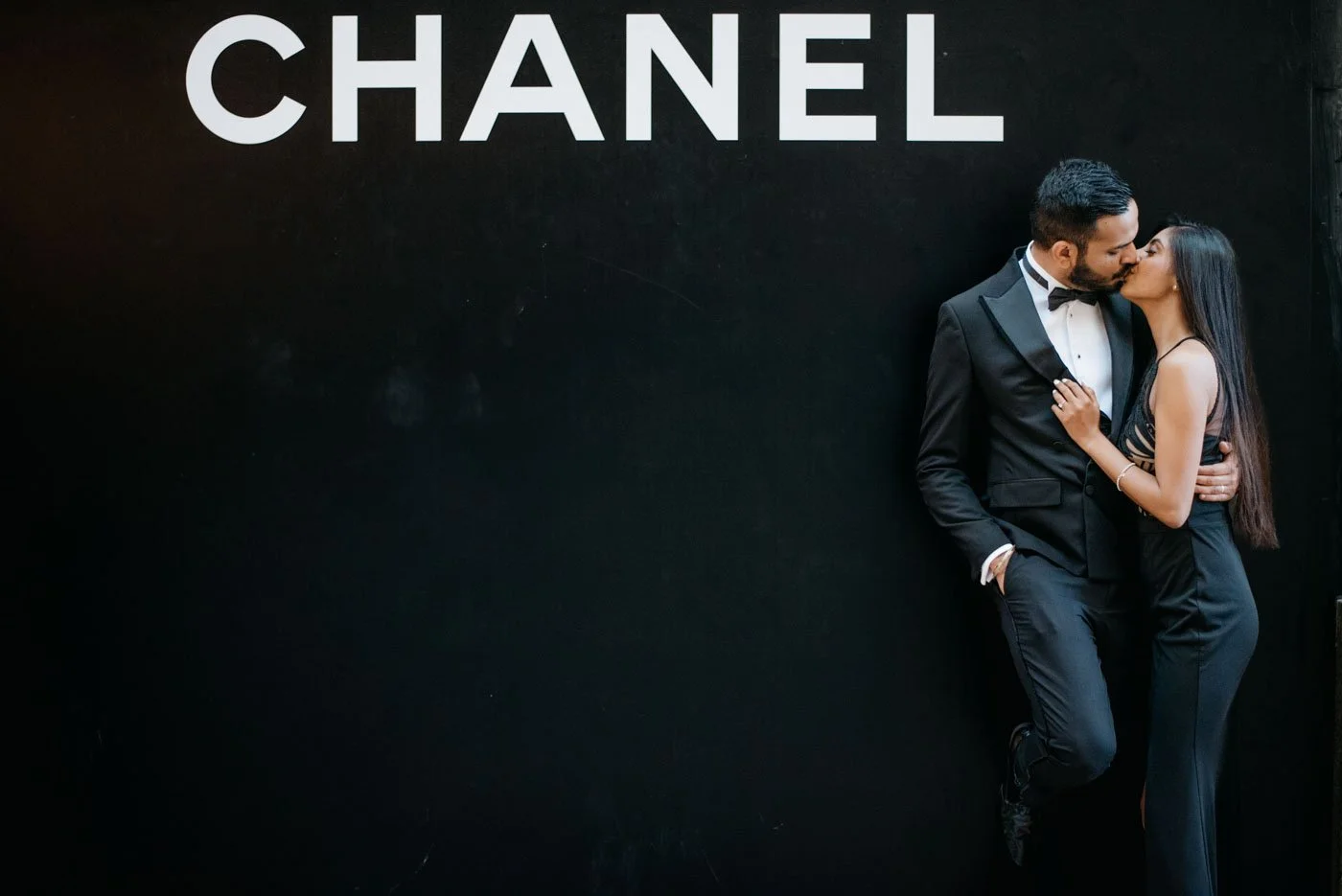 A couple in formal attire kissing against a black wall with the word 'CHANEL' written on it.