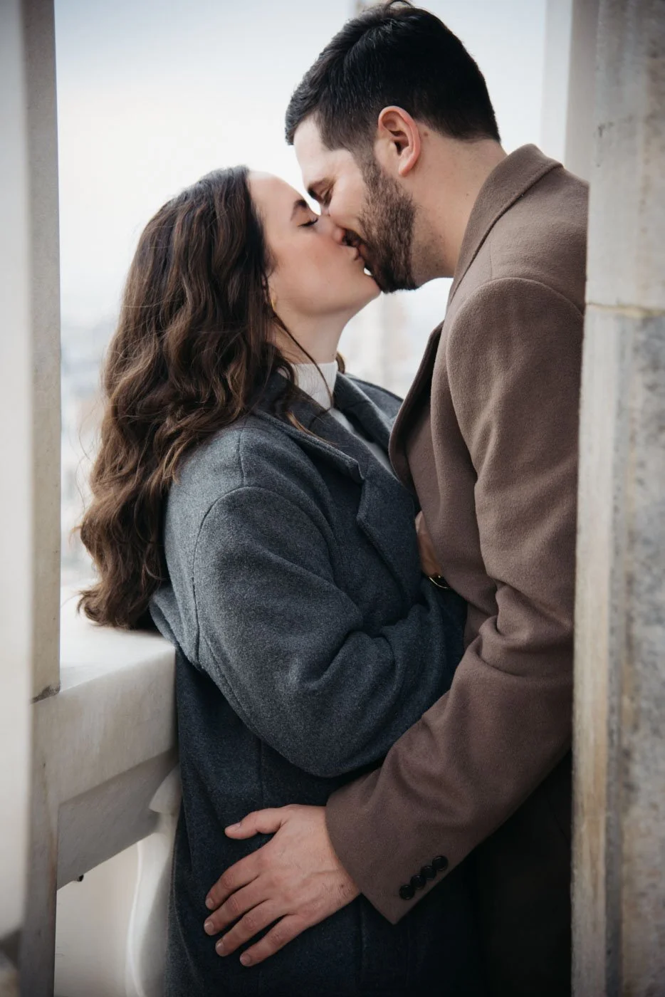 A man and woman are kissing between two stone columns, standing close with eyes closed, on a building balcony with cityscape in the background.