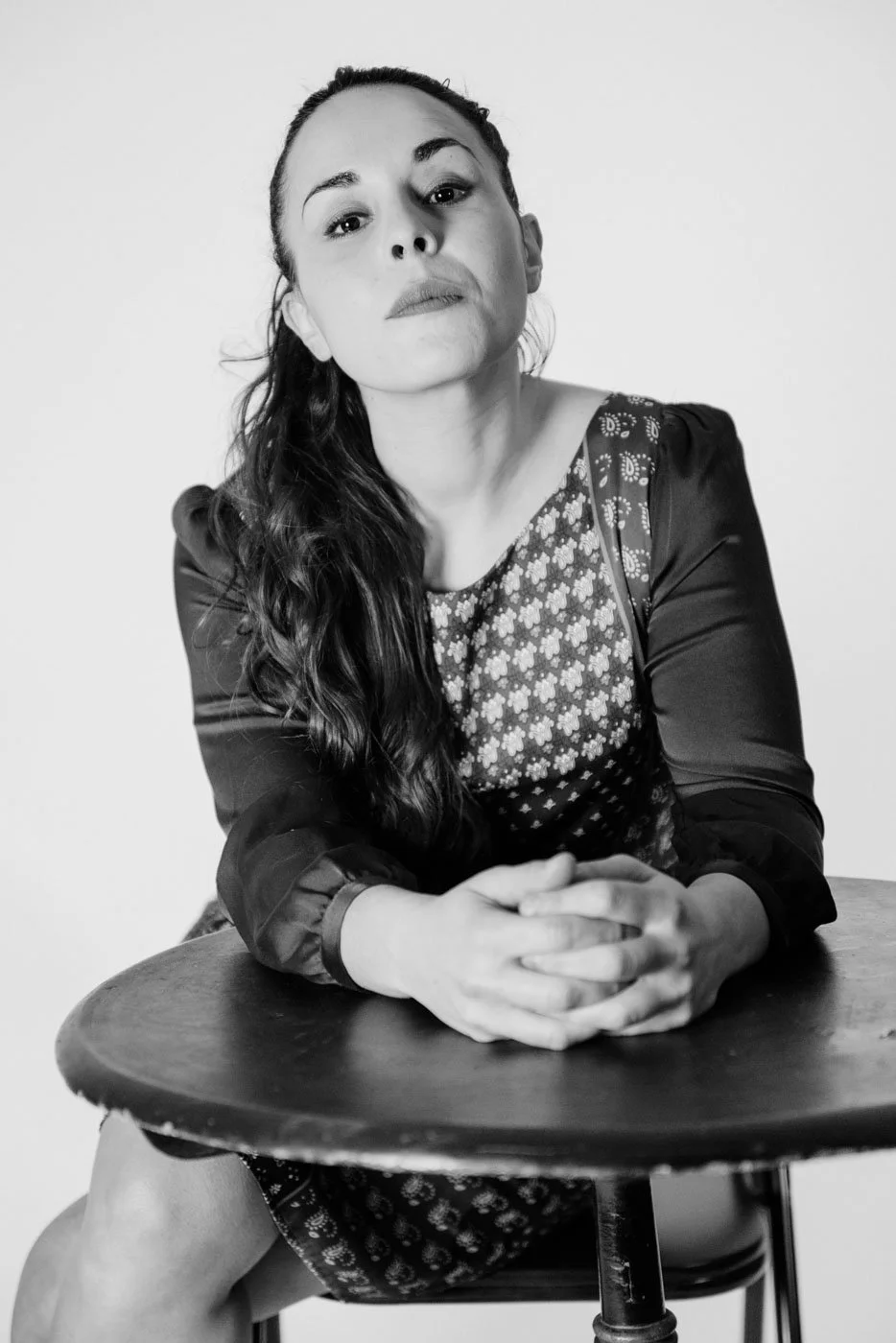 Black and white photo of a woman with long wavy hair sitting at a table, resting her arms on the table with her hands clasped.