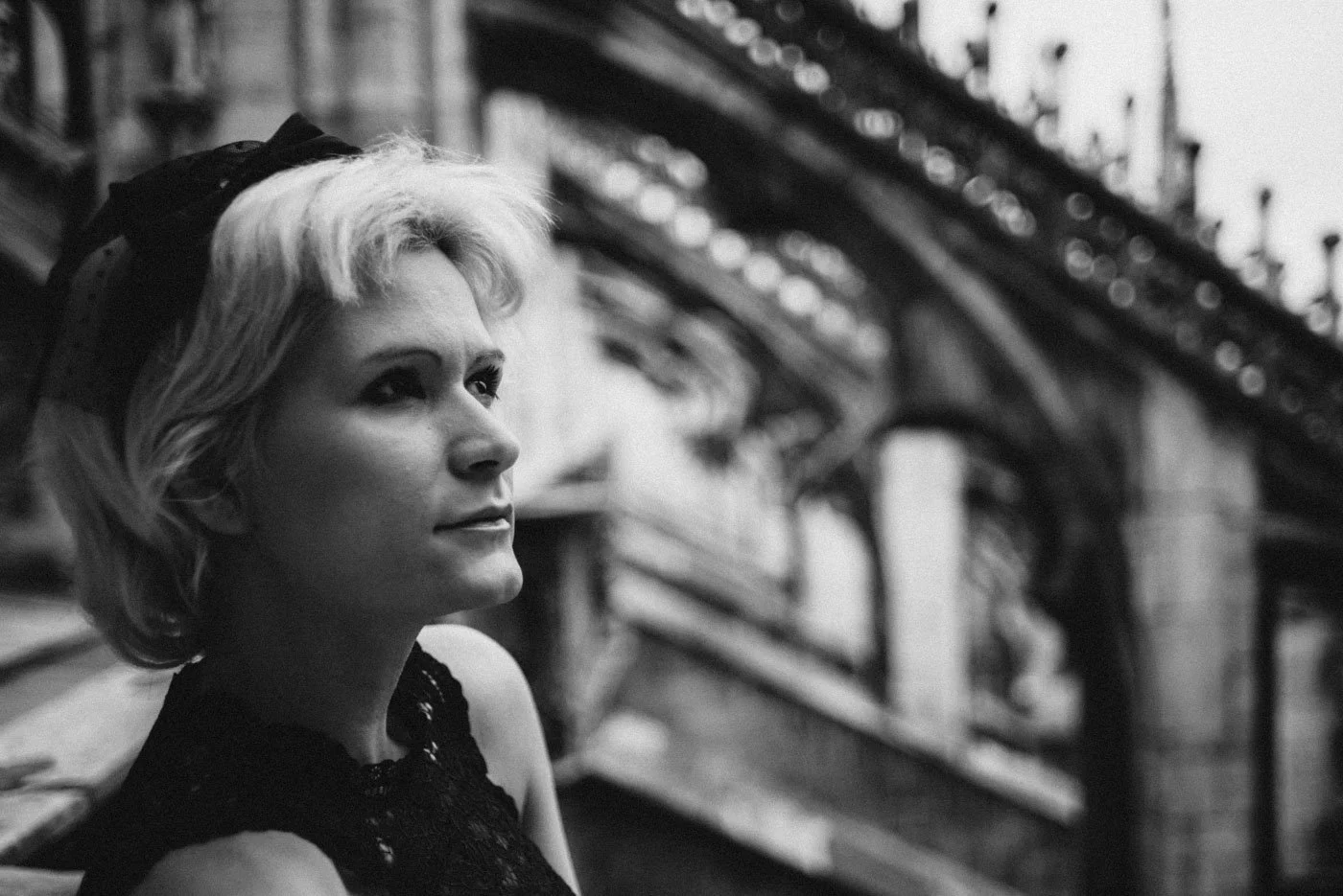 A woman with short blonde hair sitting on a bench under a bridge, looking thoughtfully into the distance, in black and white.