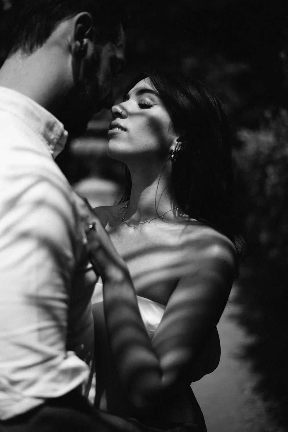 A black and white photo of a man and woman close to each other, with their faces inches apart, sharing an intimate moment outdoors at night.