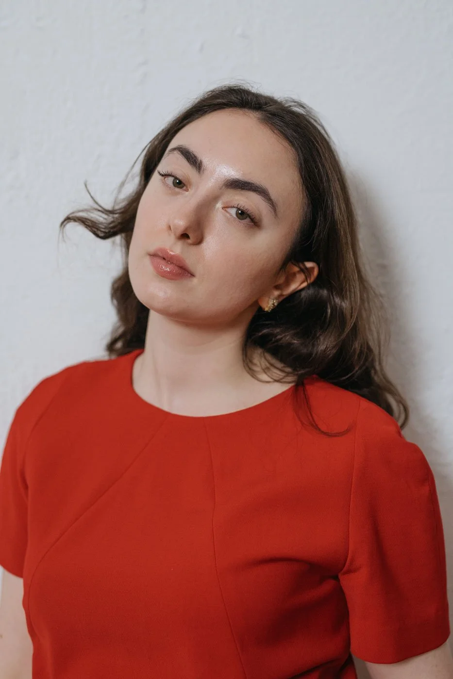 A young woman with wavy brown hair wearing a red top, standing against a white wall, looking into the camera with a neutral expression.