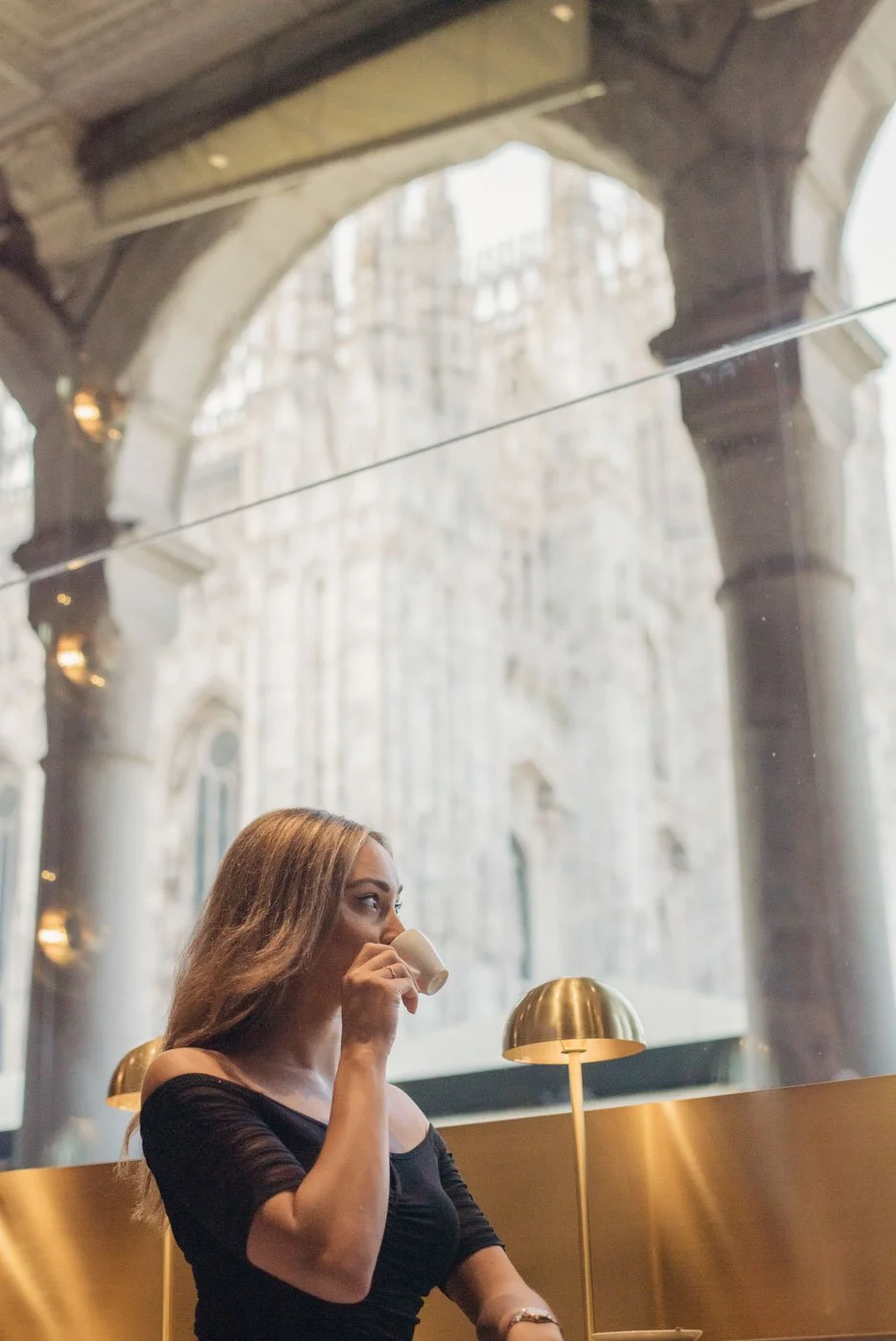A woman with long brown hair drinking from a white cup in a modern café with gold lamps and large arched windows, with a view of a historic building outside.