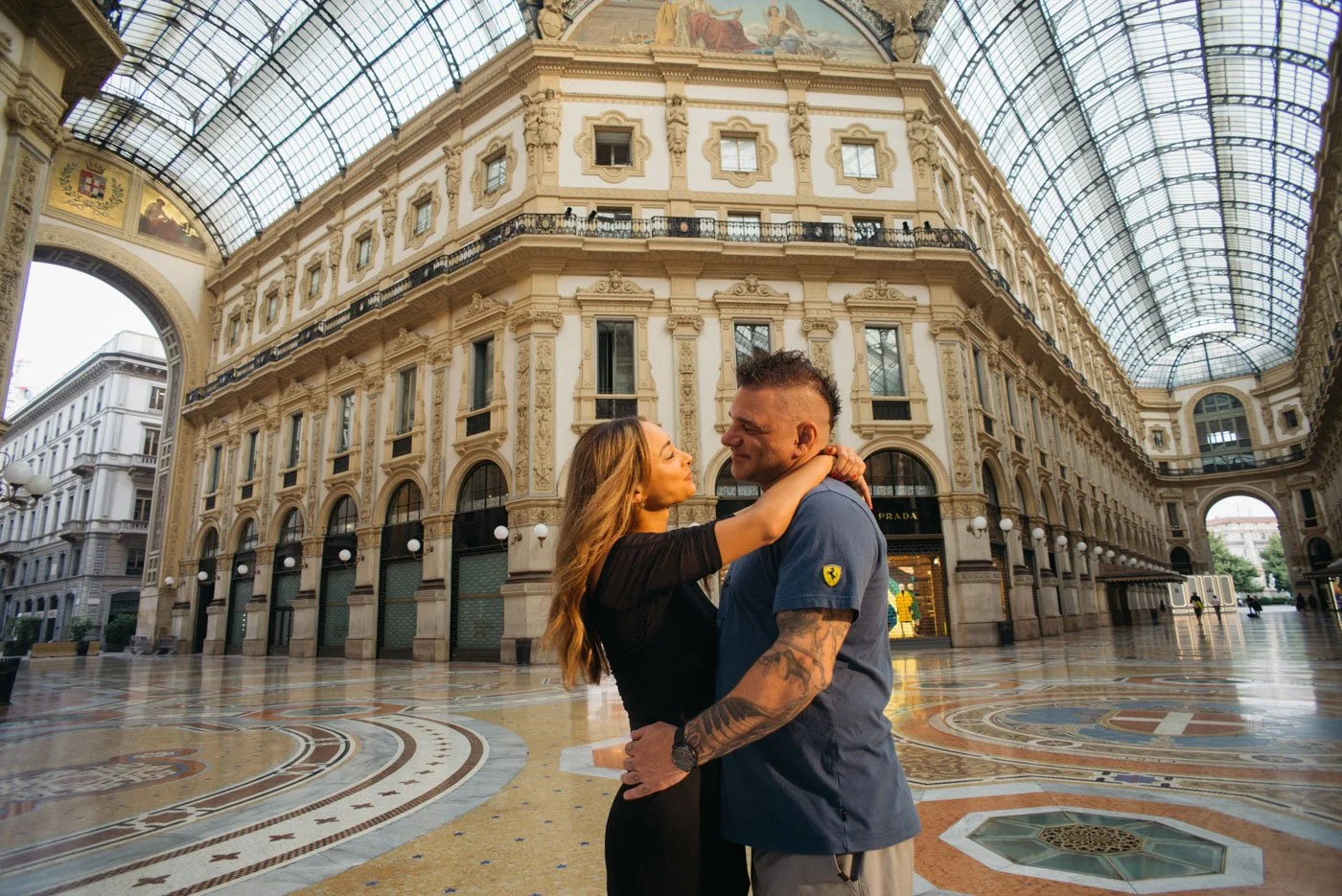A couple embracing and smiling in a grand, historic shopping arcade with ornate architecture and a glass ceiling.