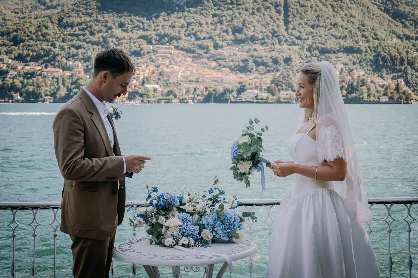 Bride and groom exchanging vows on a lakeside balcony with a scenic mountain background, flowers on a table between them.