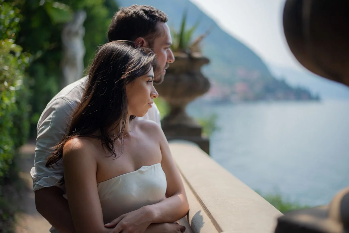 A man and woman sit closely together outdoors near water, with mountains in the background, looking into the distance.