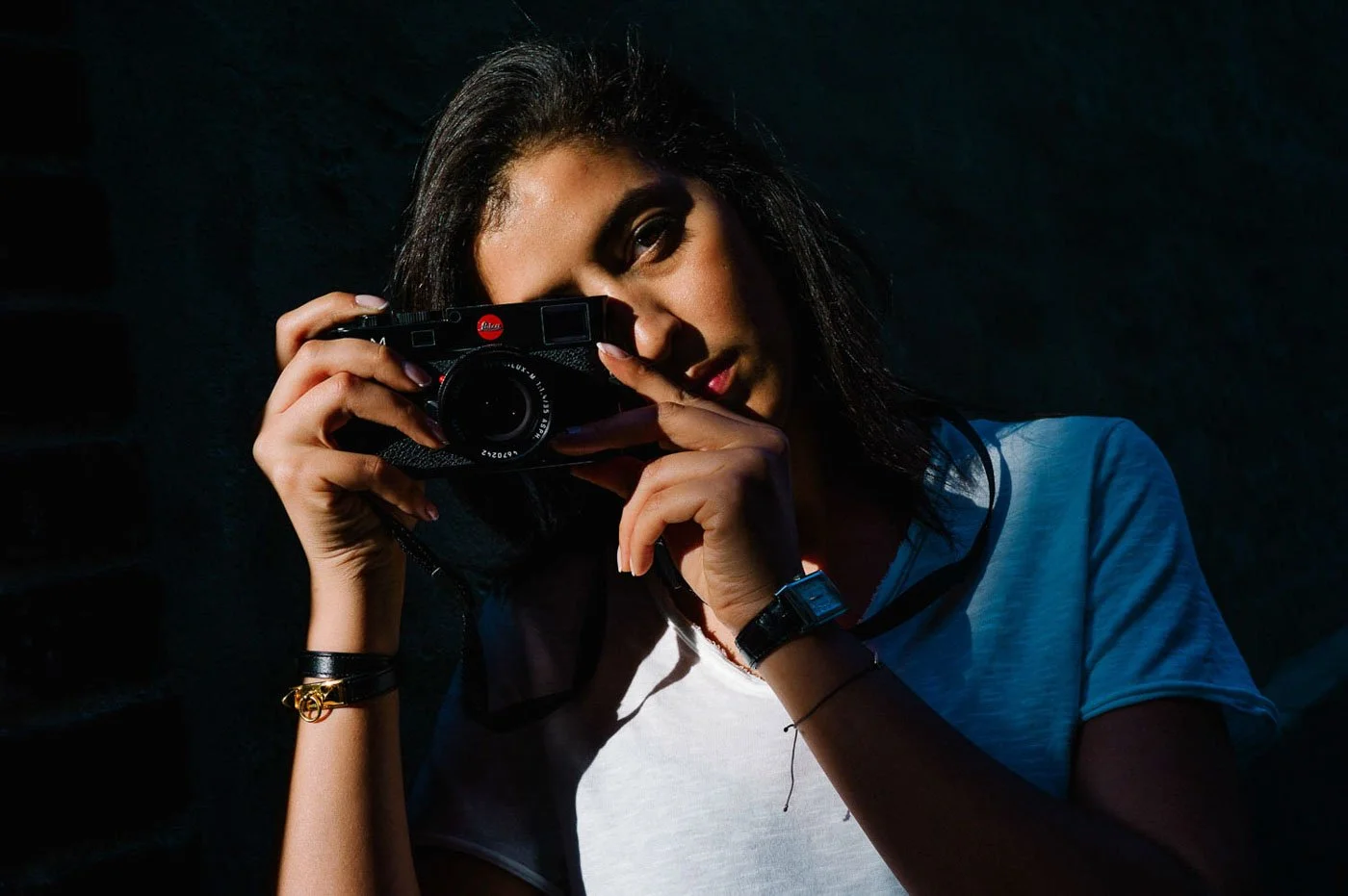 Woman with dark hair holding a camera up to her face, wearing a light blue shirt and a watch, in a dark setting