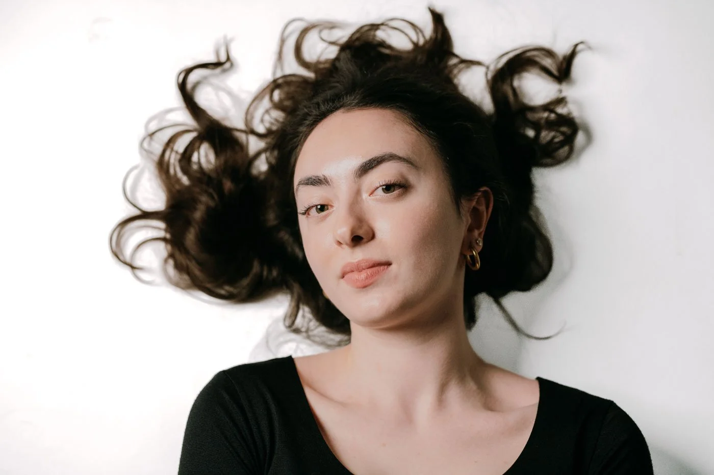 A young woman with dark, wavy hair lying on a white surface, wearing a black top and hoop earrings.