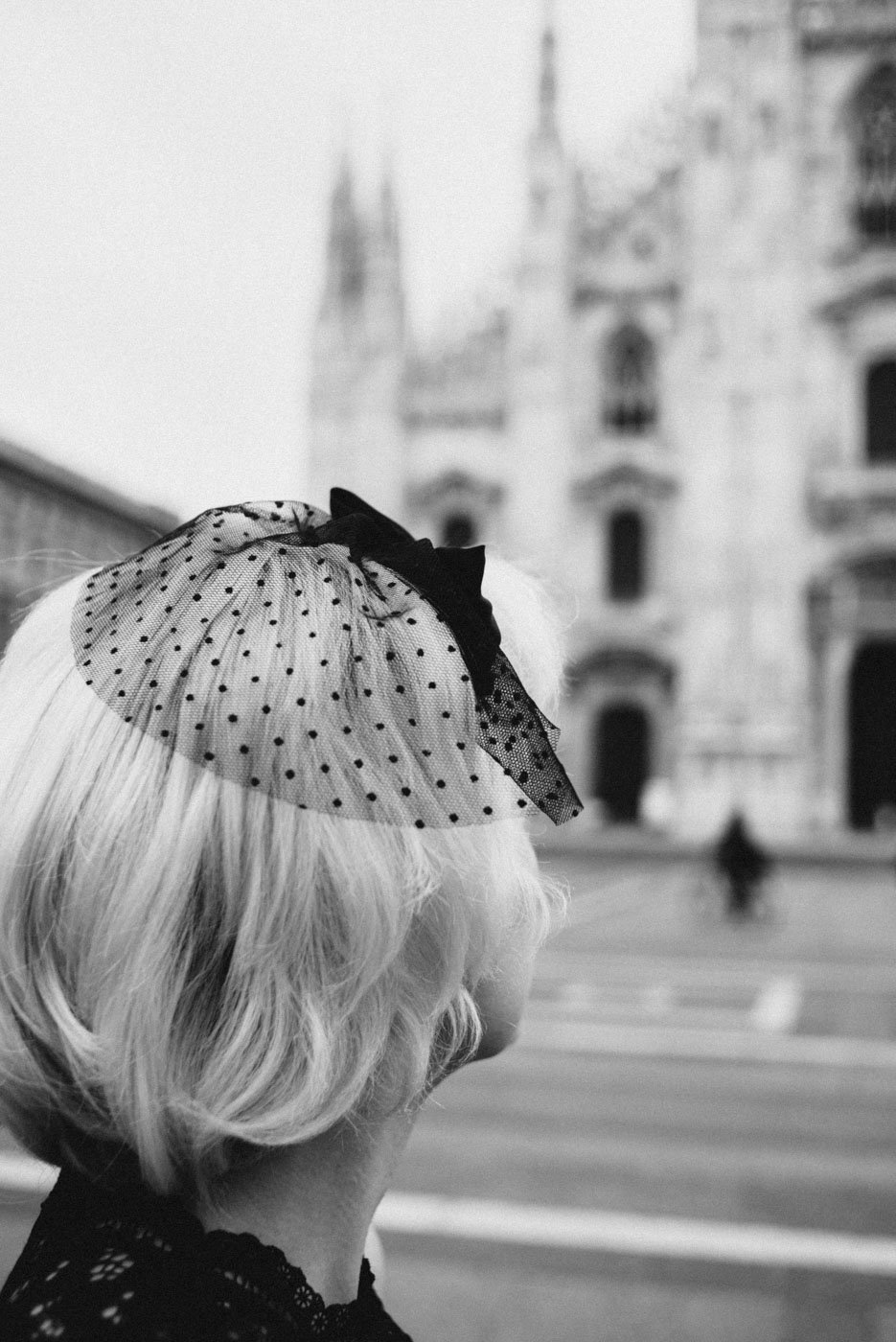A black-and-white photo of a woman with short, wavy blonde hair, wearing a veil with polka dots and a bow, looking at a historic European building with tall towers in the background.