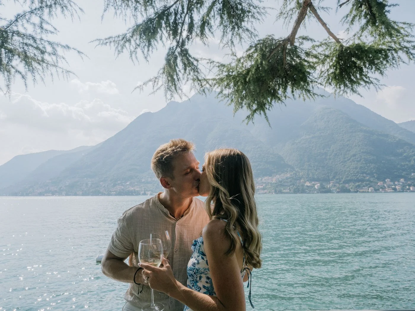 A couple sharing a kiss with a lake and mountains in the background, with overhanging tree branches at the top.