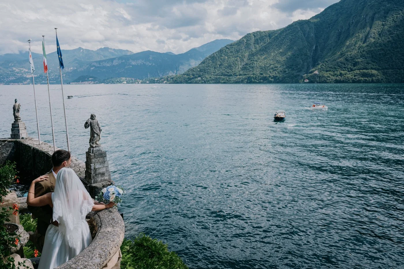 A bride and groom standing on a stone balcony overlooking a lake with boats and mountains in the background, during a wedding ceremony.