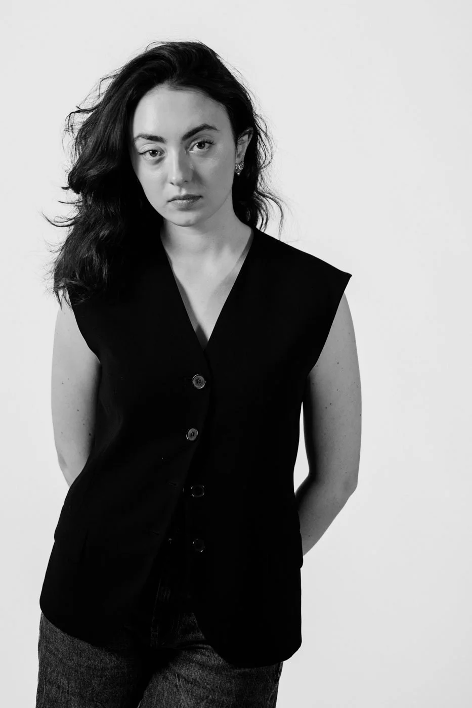 Black and white portrait of a woman with wavy dark hair, wearing a sleeveless black vest with buttons, standing against a plain background.