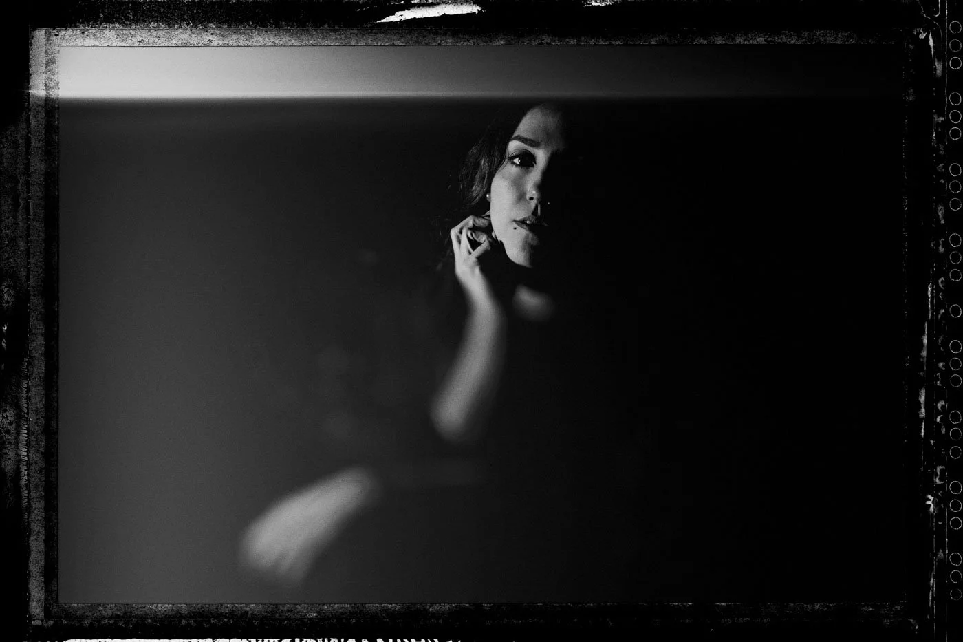 Black and white close-up portrait of a woman with dark hair, looking at the camera with her hand near her face, partially illuminated with shadows creating a dramatic effect.