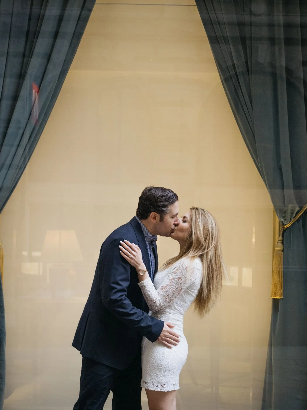A couple sharing a kiss behind a glass window with dark curtains on each side, in a warmly lit indoor setting.