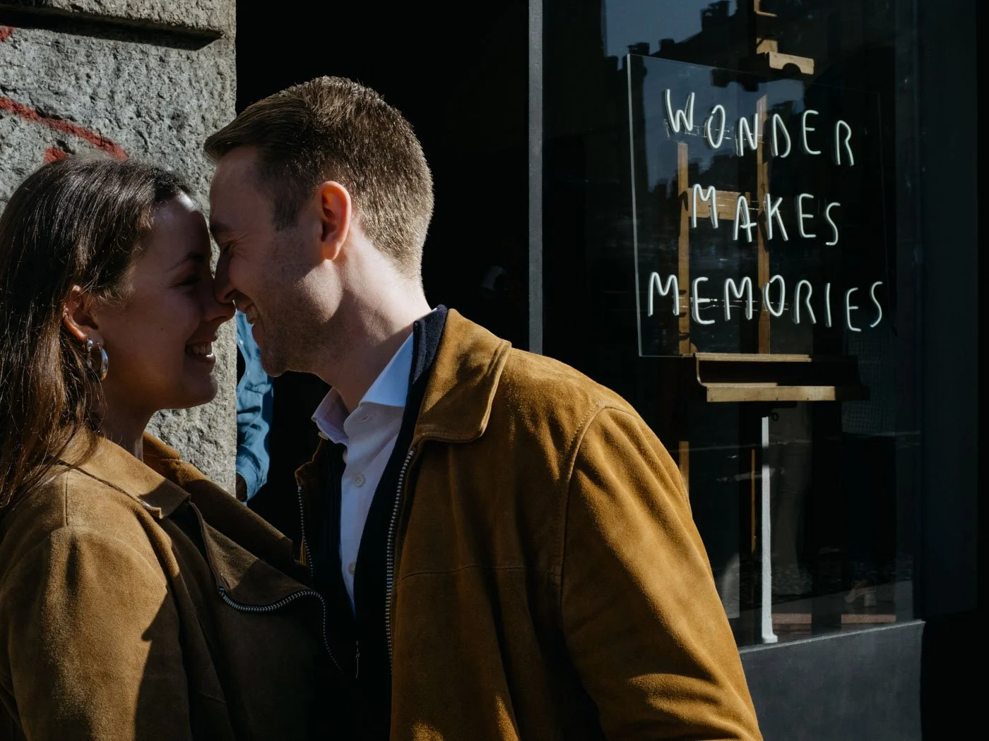 A couple is smiling and touching foreheads outside a building with a sign that reads 'Wonder Makes Memories'.