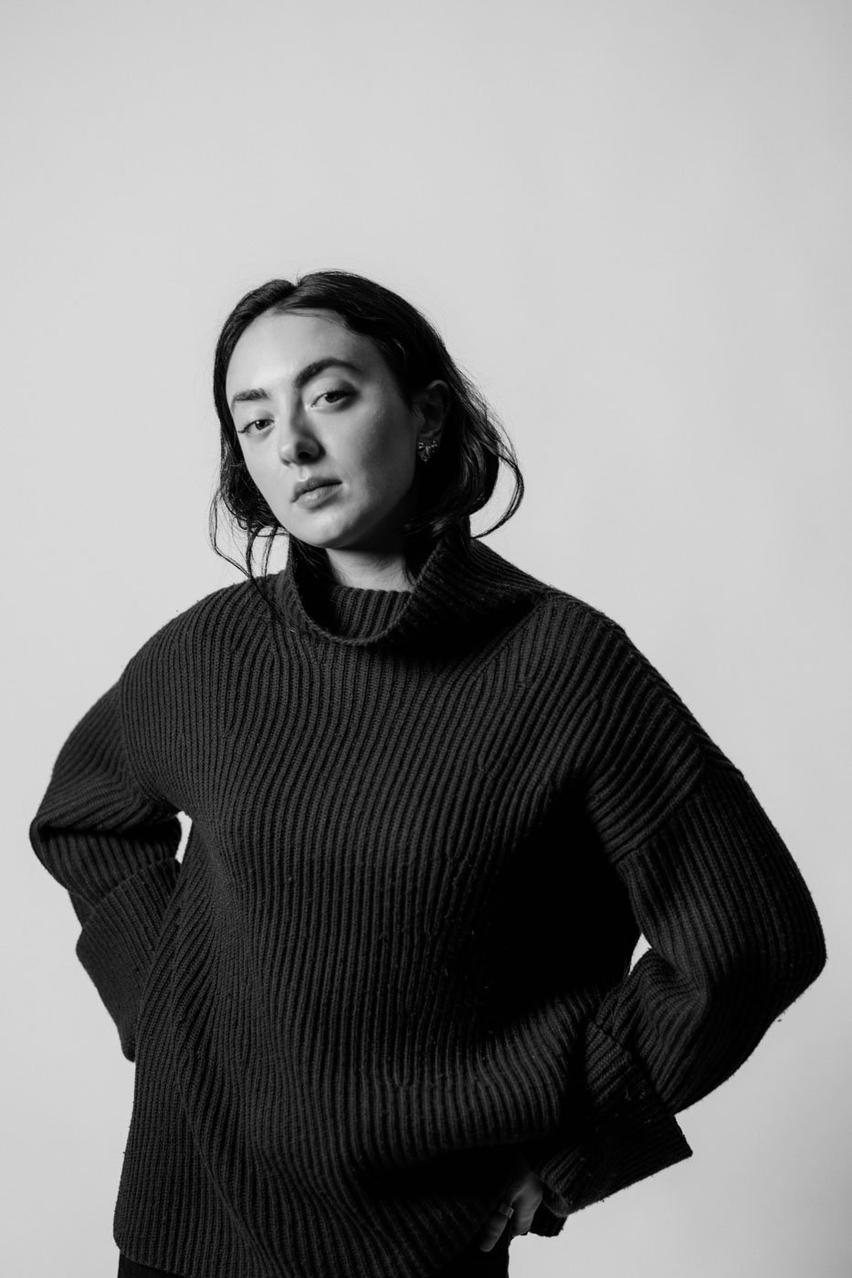 Black and white portrait of a young woman with dark hair, wearing a ribbed sweater, standing against a plain background with a confident expression.