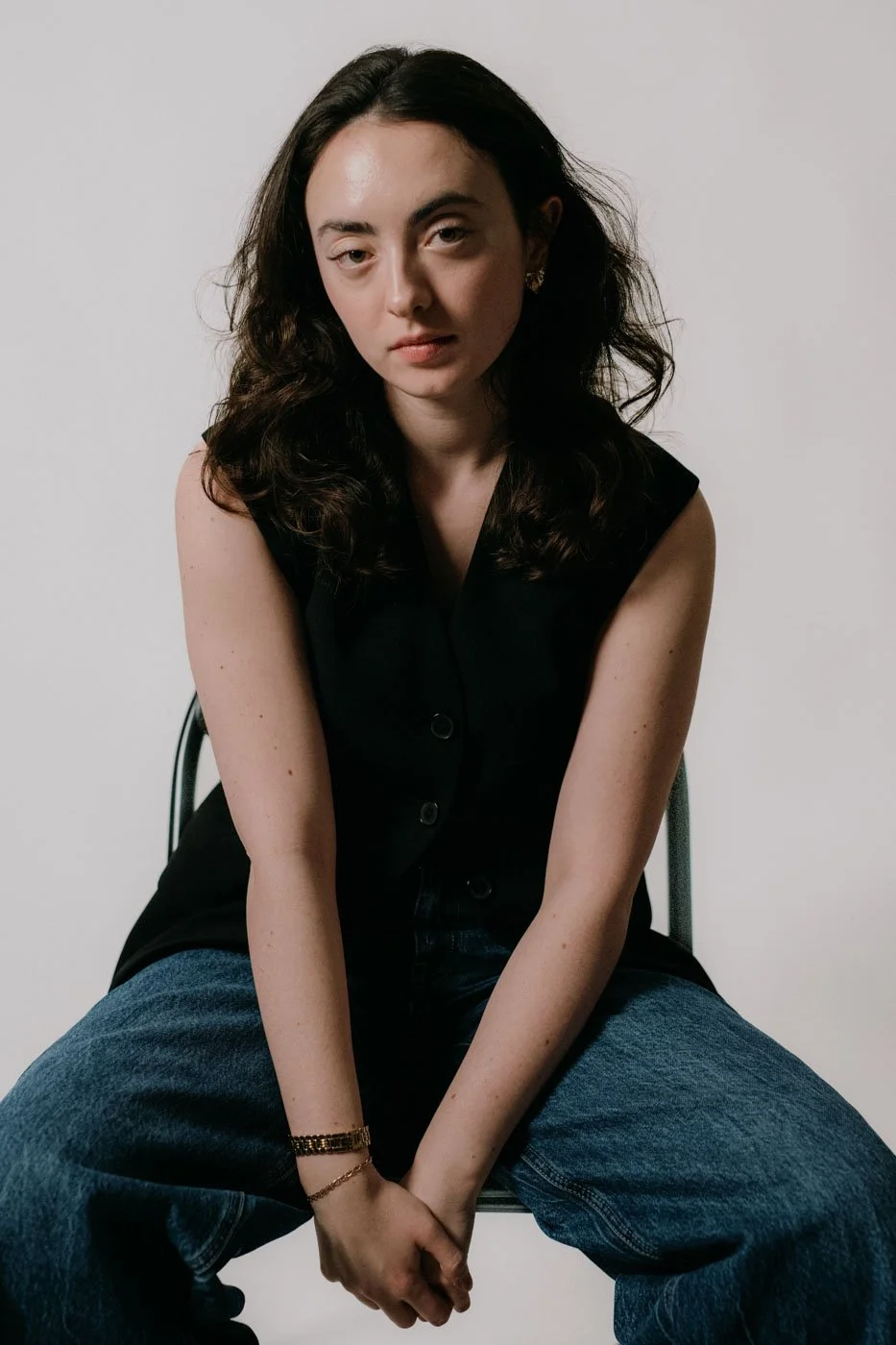 A young woman with curly dark hair, wearing a sleeveless black top and blue jeans, sitting on a chair against a neutral background.
