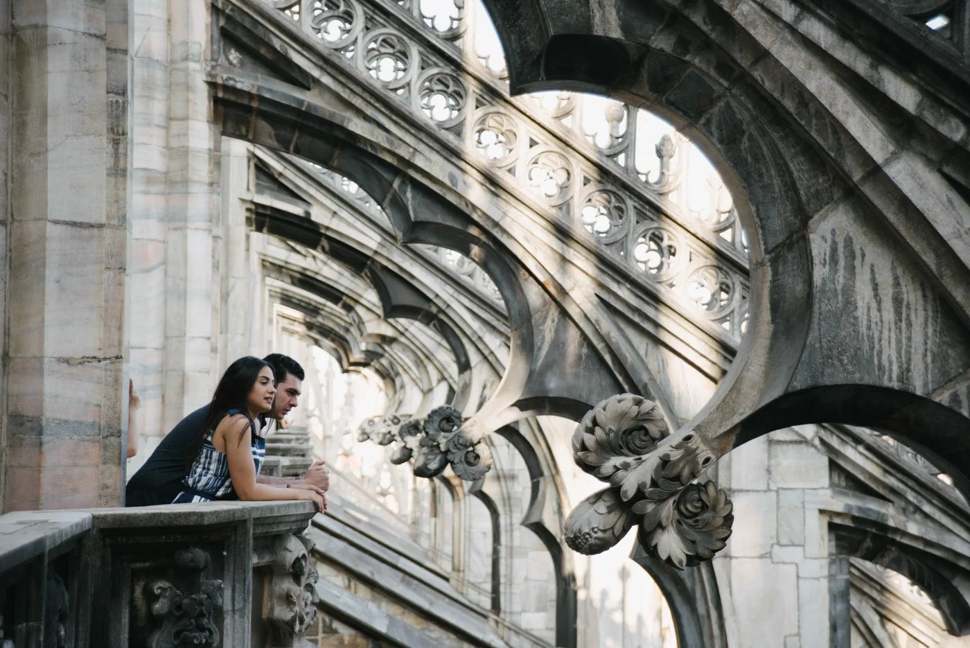 Three people, two women and one man, leaning over a stone balcony railing, looking at ornate architectural details on a historic building with Gothic-style arches and floral carvings.