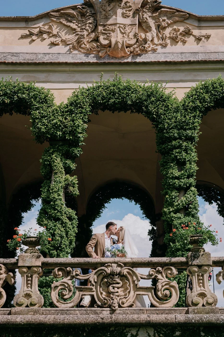 A newlywed couple sharing a kiss on a decorative stone balcony with lush greenery and architectural details.