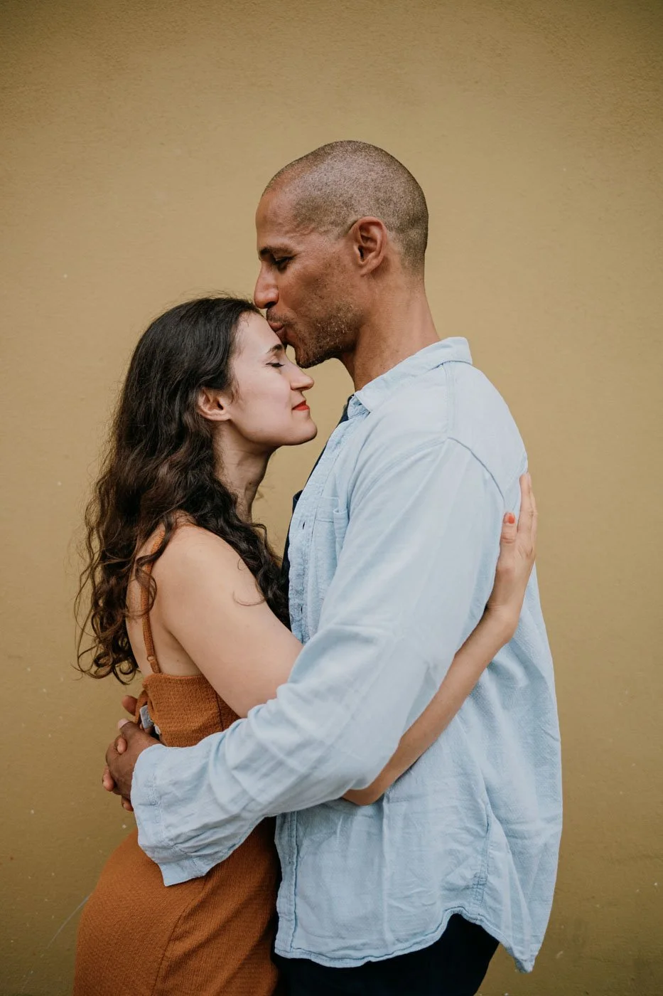 A man with a shaved head gently kisses a woman with curly dark hair on her forehead as they embrace against a plain beige wall.