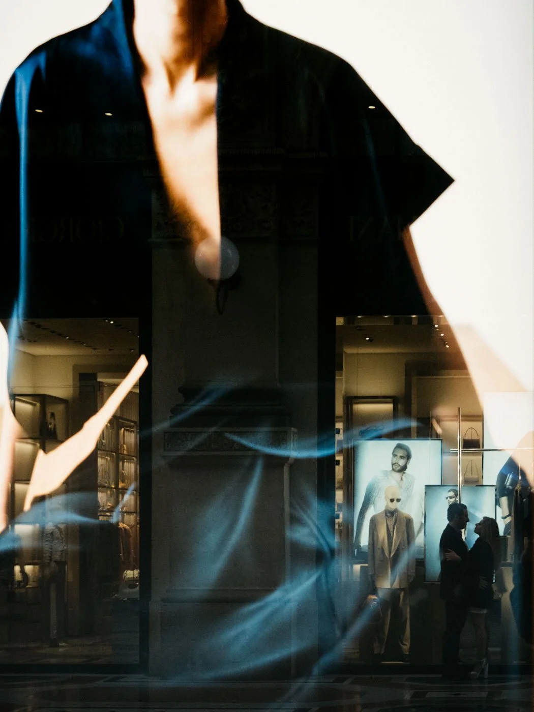 Reflection of a mall storefront window with mannequins and large posters of male models inside, with two people talking in the background.