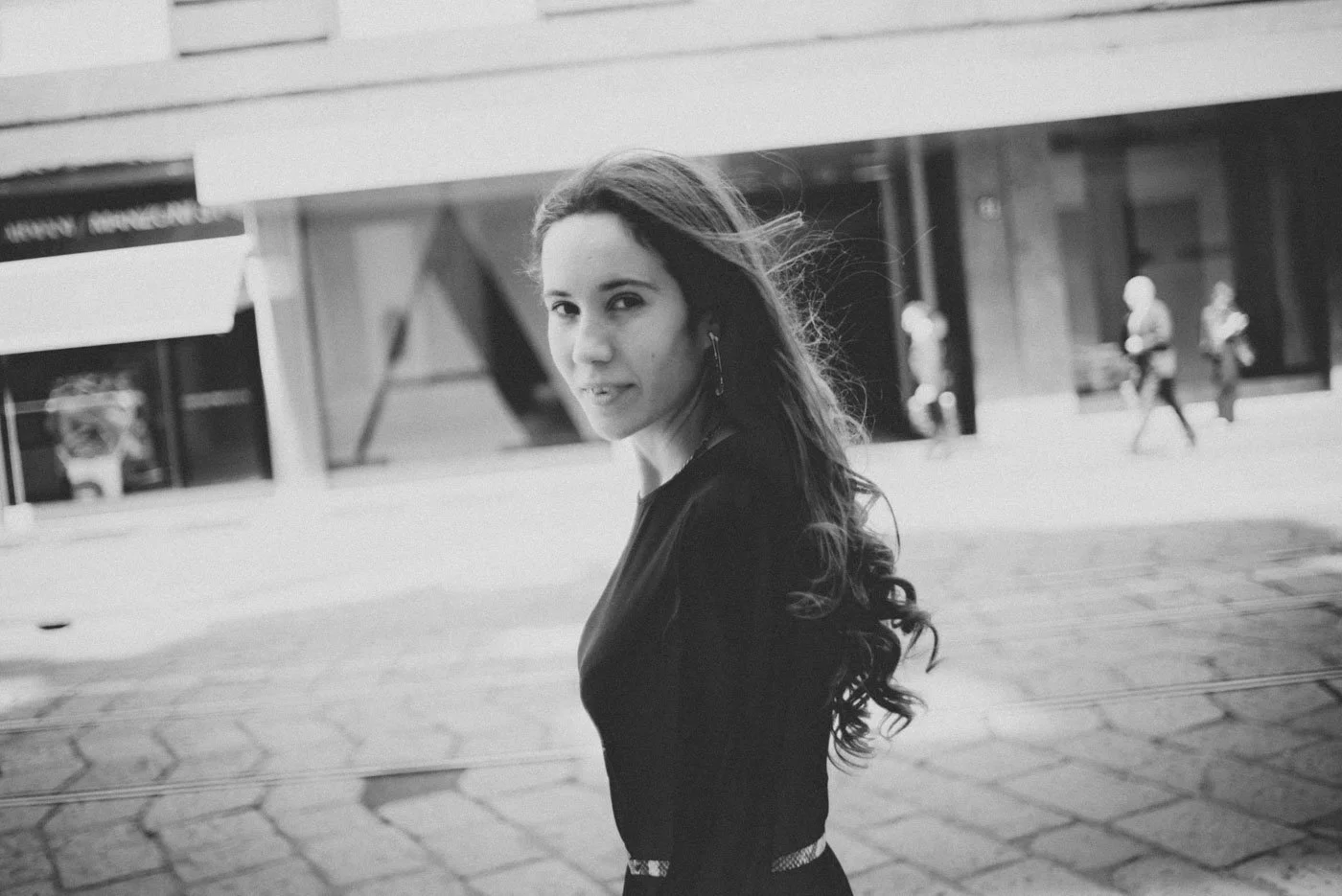 Black and white photo of a woman with long wavy hair looking back over her shoulder outside in an urban area with buildings and pedestrians in the background.