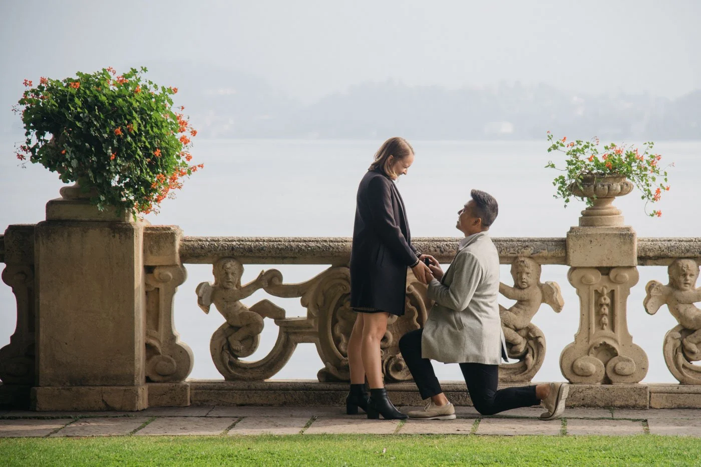 A man proposes to a woman by kneeling and holding her hand on a stone balcony overlooking a body of water, with potted flowers on either side.