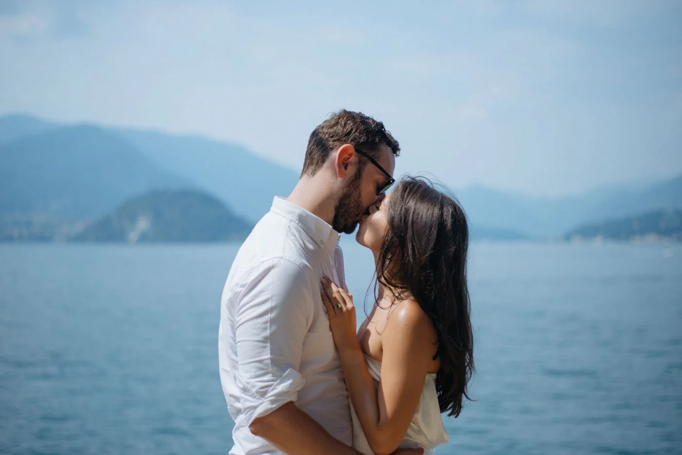 A couple sharing a kiss by a lake with mountains in the background on a clear day.