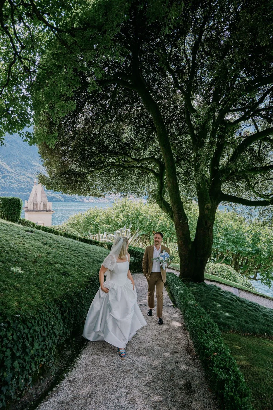 Bride and groom walking on a garden pathway near a lake, surrounded by greenery and a large tree, with a church steeple in the background.
