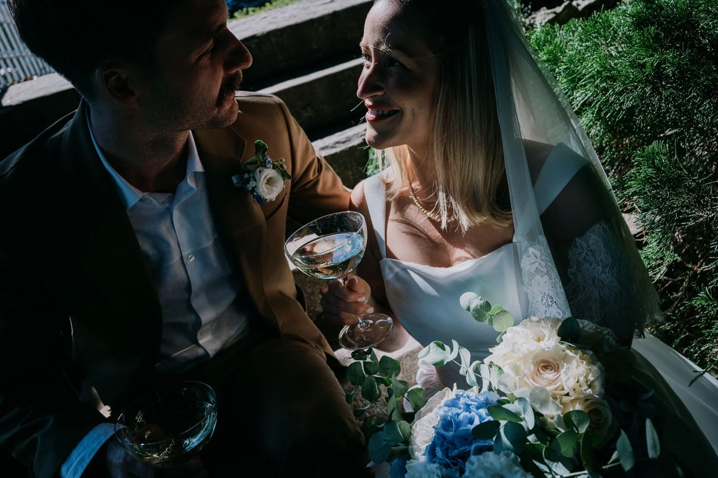 A bride and groom sit closely, smiling at each other during their wedding, with the bride holding a bouquet of white and blue flowers.