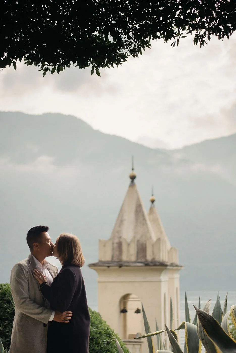 A couple kissing outdoors near a church steeple with mountains in the background.