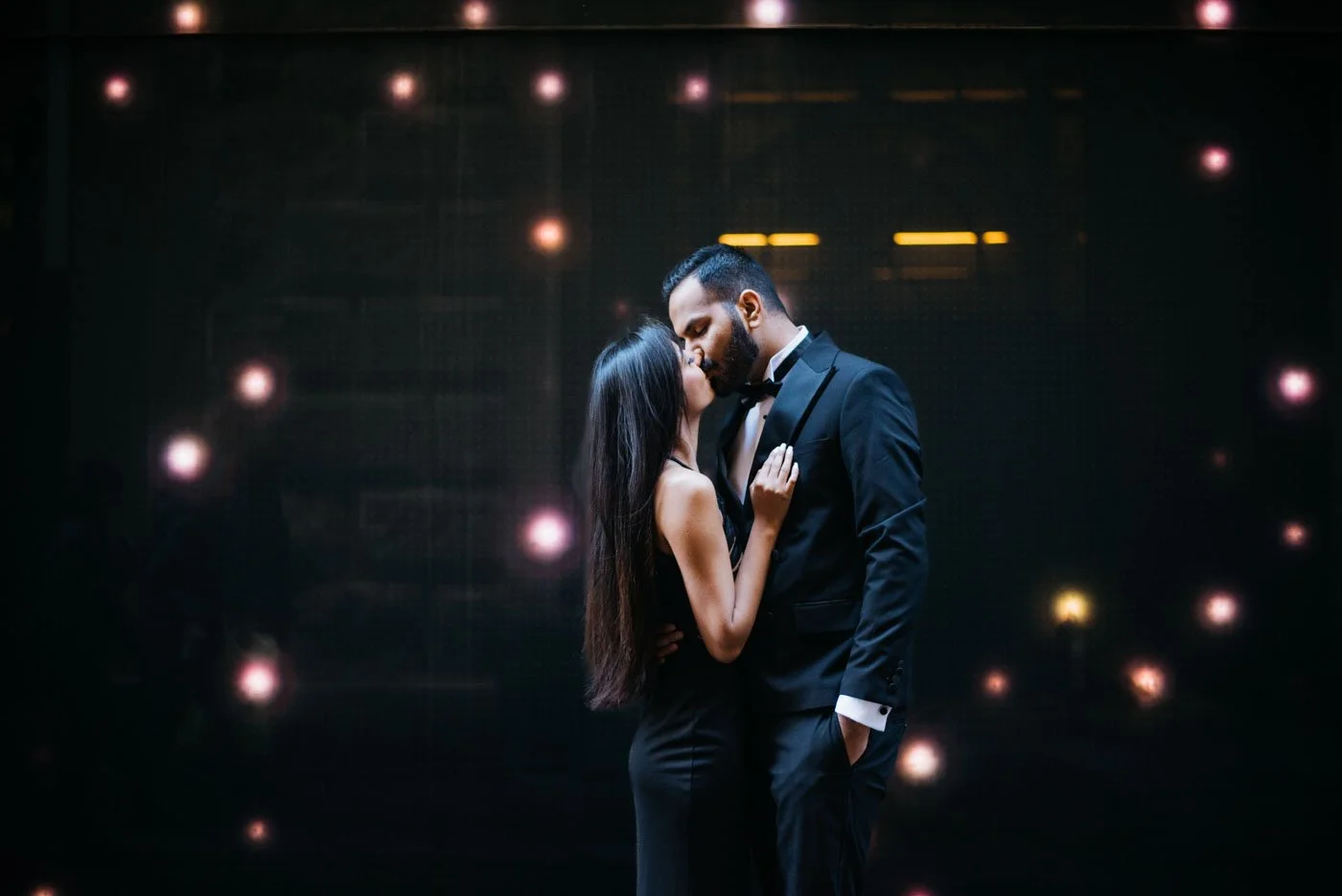 A couple in formal attire sharing a kiss, with the man in a tuxedo and the woman in a black dress, against a dark background with soft, out-of-focus lights.