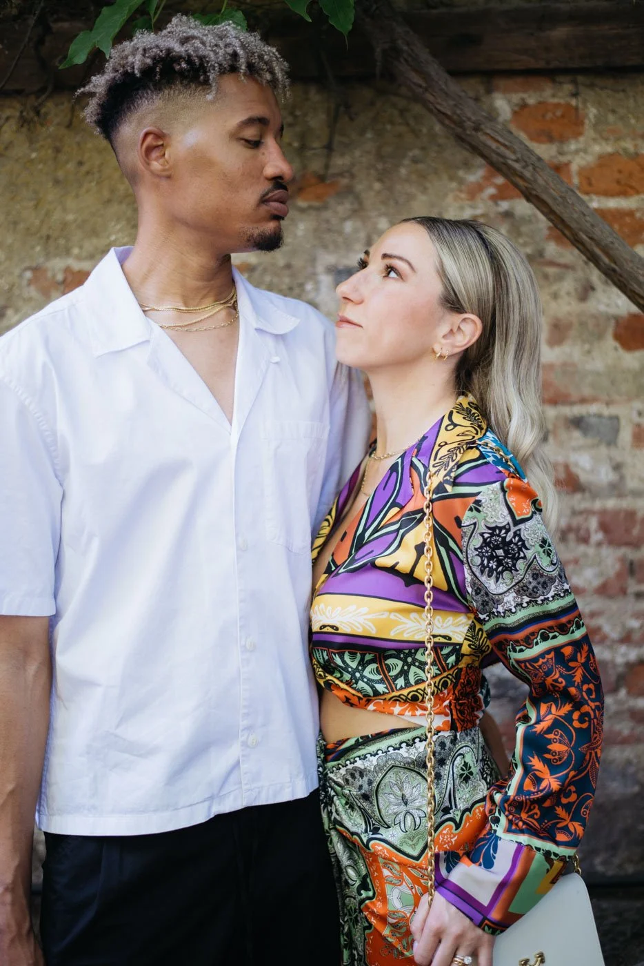 A man and a woman standing close together outdoors, looking into each other's eyes, with a rustic brick wall and wooden beam in the background. The man is wearing a white button-up shirt and gold necklaces, while the woman is dressed in a colorful, p