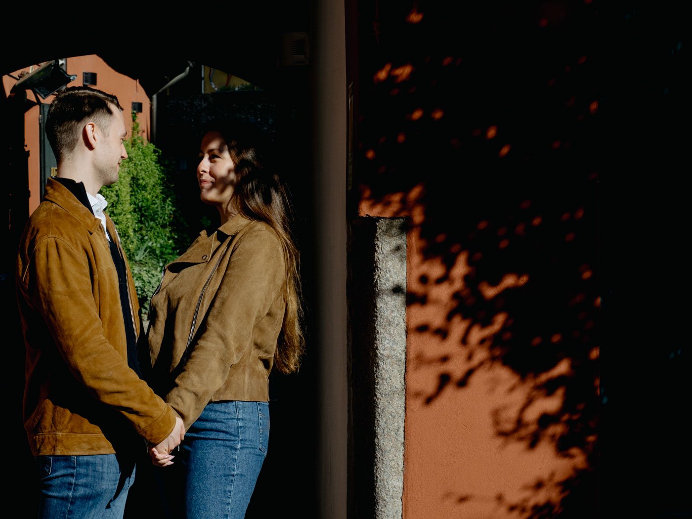 A young couple standing face to face, holding hands, looking into each other's eyes outdoors during daytime, with shadows of trees falling on a wall behind them.