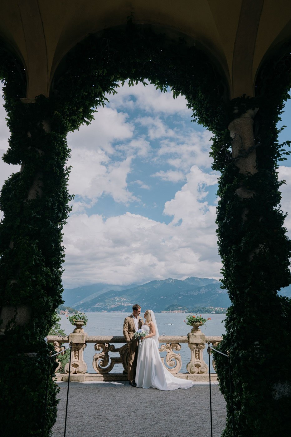 A bride and groom standing on a terrace with a lake and mountains in the background, framed by an archway covered in greenery.