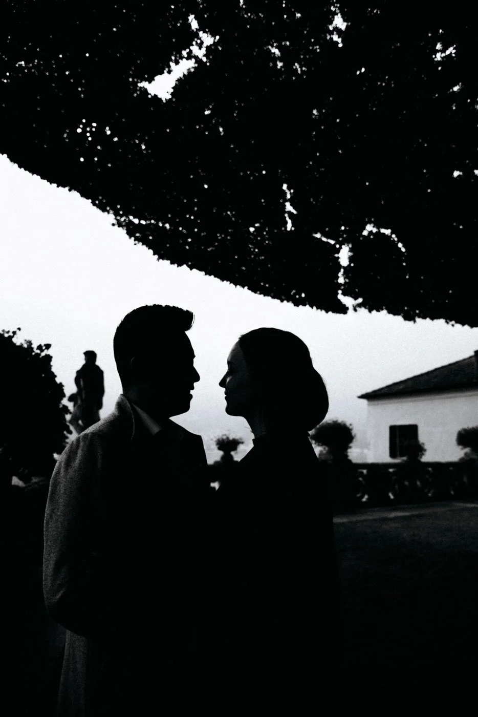 Silhouettes of a man and woman facing each other closely outdoors during daytime with a tree canopy overhead, a house, and other people in the background.