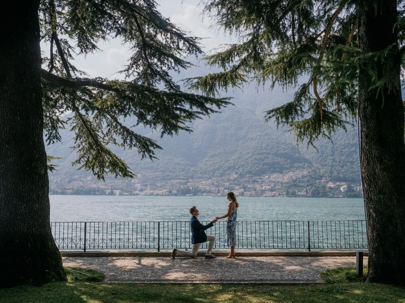 A man proposing to a woman by a lake, framed by two large trees, with mountains and a town in the background.