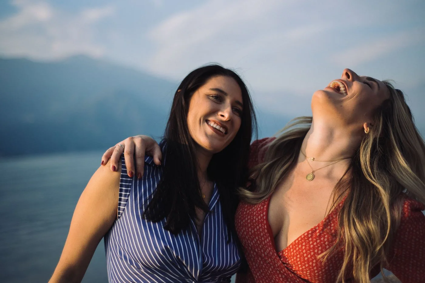 Two women smiling and laughing outdoors near a body of water with mountains in the background.