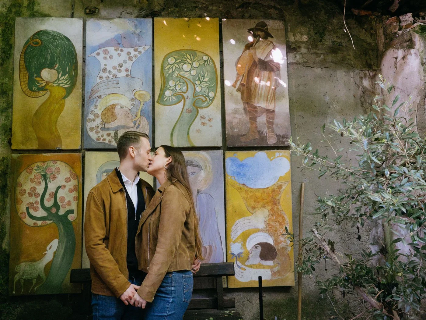 A couple kissing in front of a wall decorated with colorful paintings of trees, animals, and abstract figures, with some greenery on the right side.