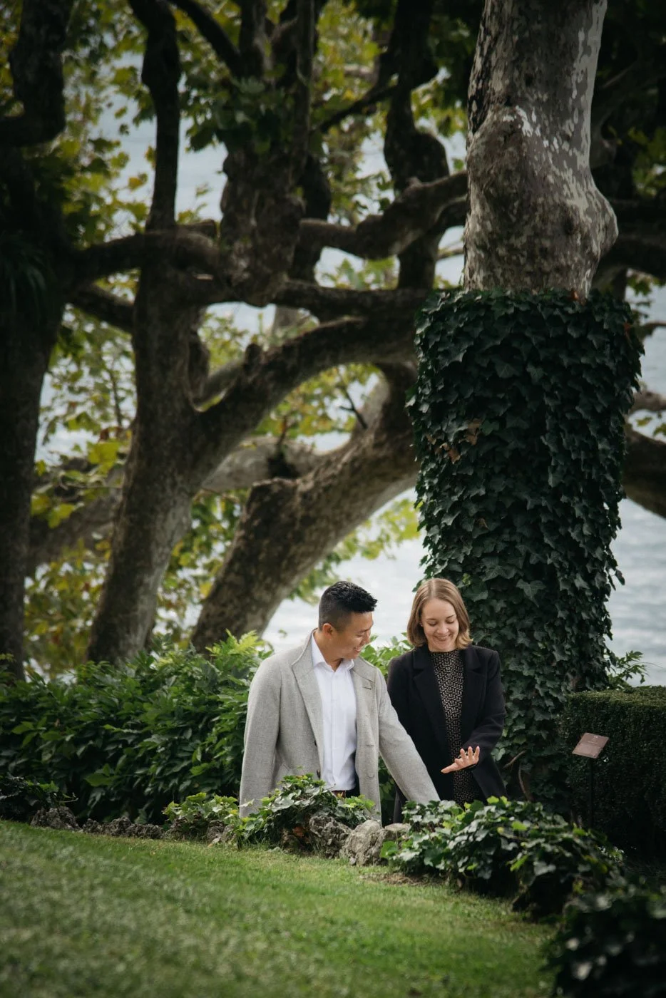Two people, a man and a woman, smiling and looking at each other outdoors near a large tree with ivy growing on its trunk. They are dressed in business casual attire, standing on a well-maintained lawn with lush plants around.
