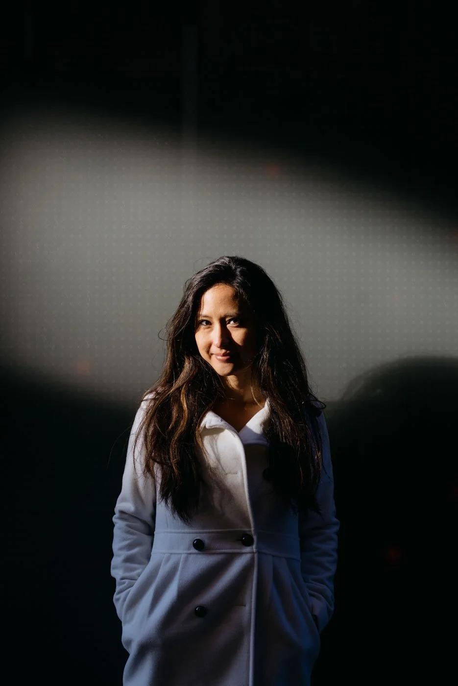 A woman with long dark hair wearing a white coat, standing in front of a dark background with a light shining on her face.
