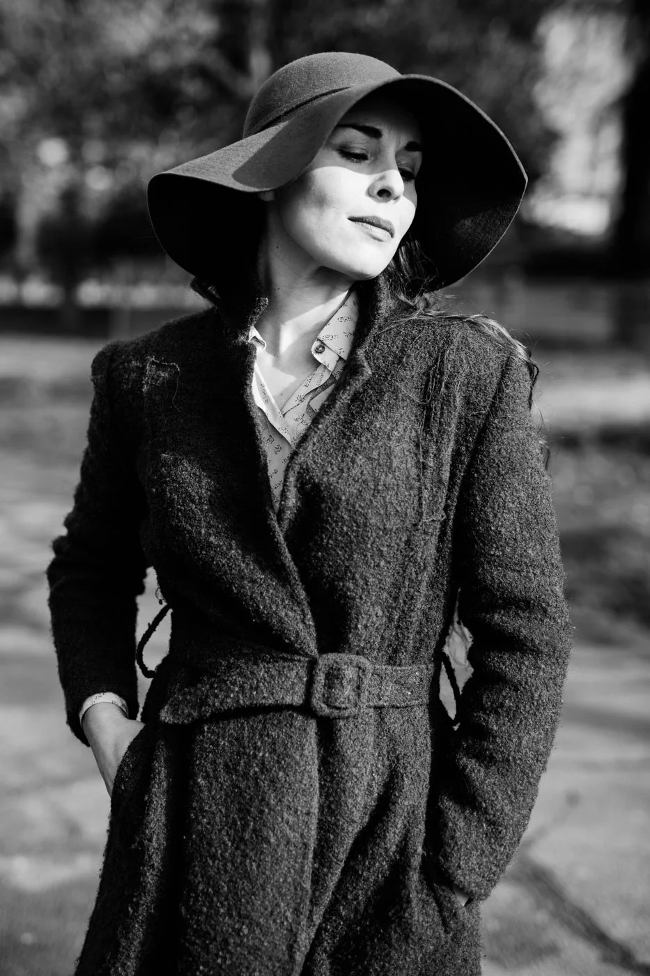 Black and white photo of a woman wearing a large brim hat and a textured coat, standing outdoors with her eyes closed.