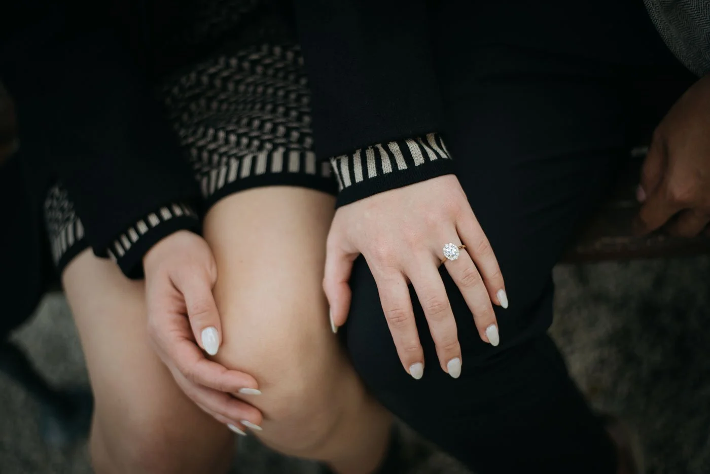 Close-up of a woman wearing a diamond engagement ring, sitting with her legs crossed, in a black outfit with patterned cuffs, resting her hand on her knee.