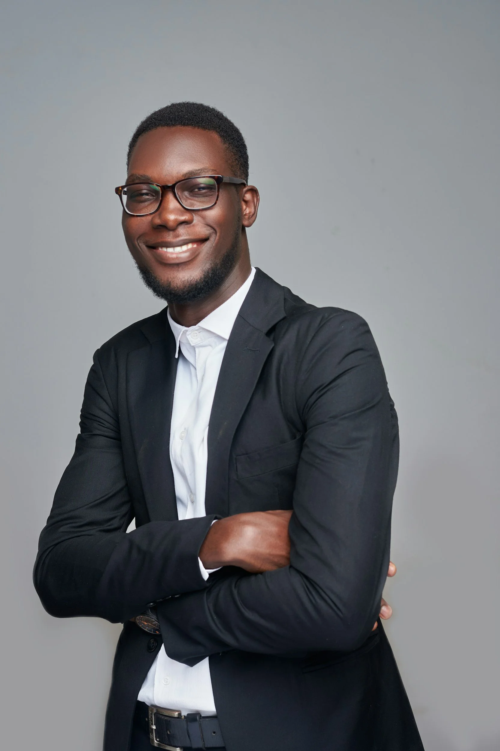 A smiling man with glasses, wearing a white shirt and black blazer, standing against a plain gray background.