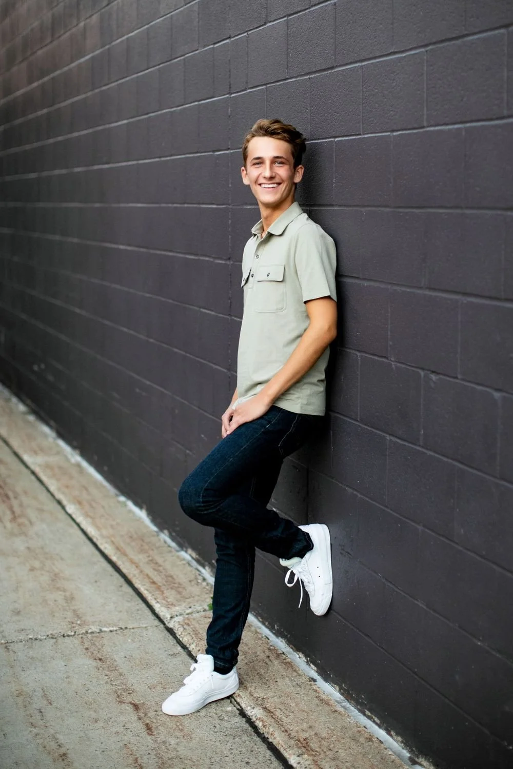 A young man with short brown hair and a big smile leaning against a dark gray brick wall. He is wearing a light green short-sleeved polo shirt, dark jeans, and white sneakers, standing with one foot slightly raised and looking at the camera.