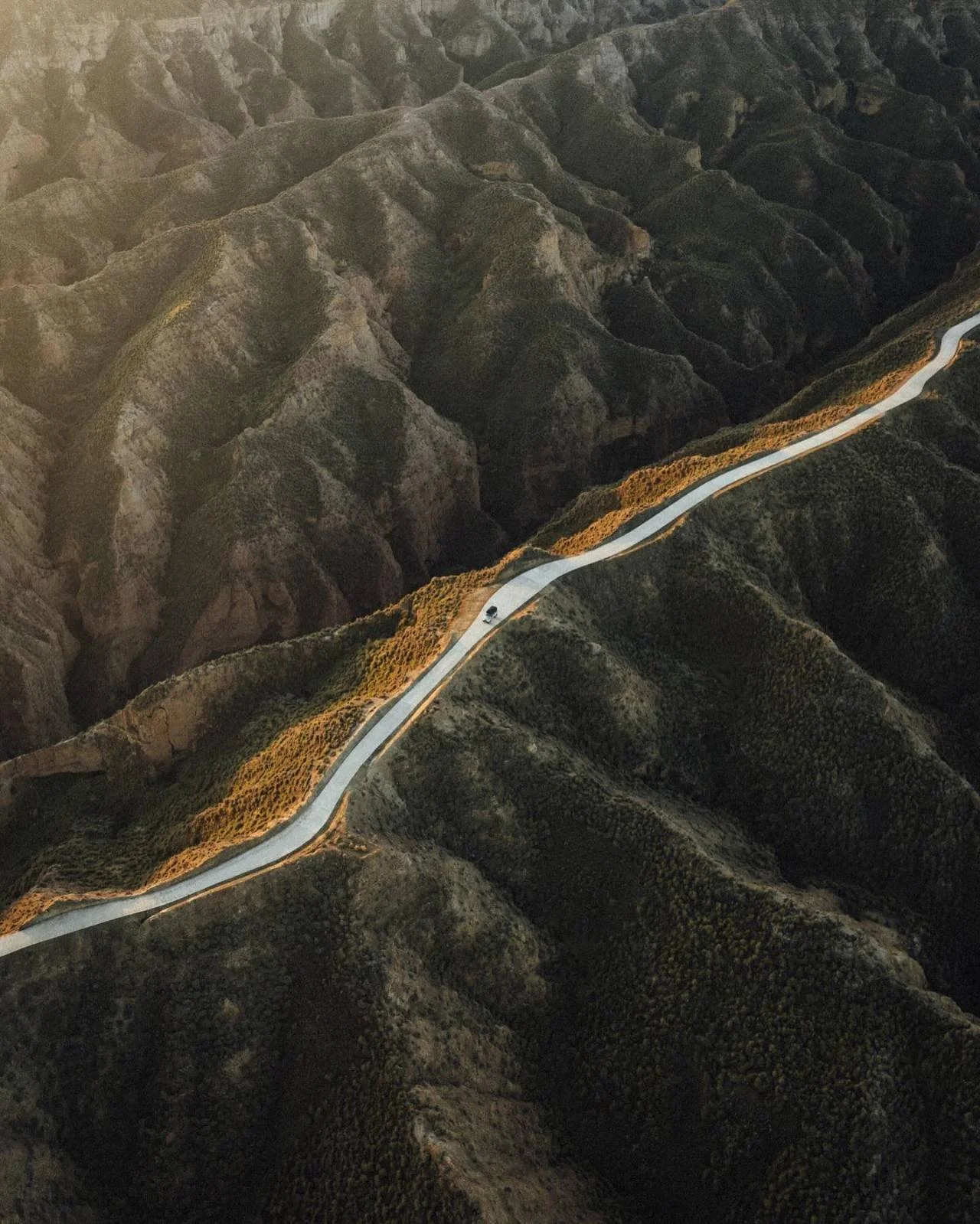 A winding mountain road with a single vehicle traveling along it, surrounded by rugged terrain and sparse vegetation.